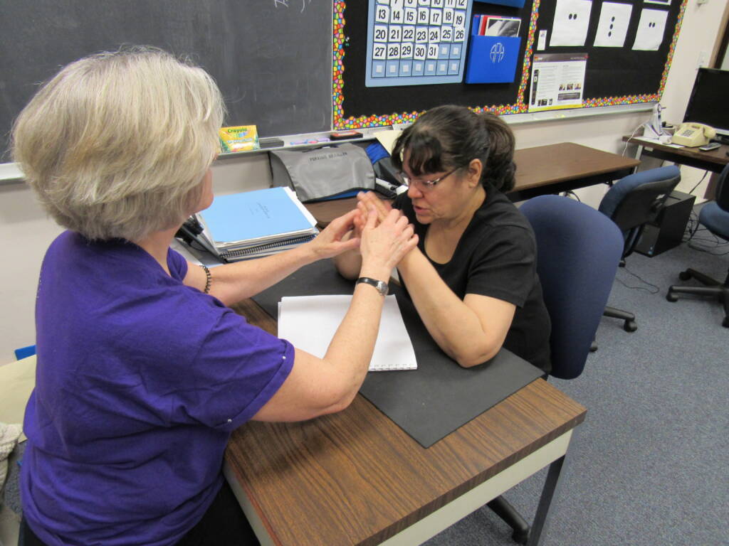 Two woman sit at a table and use tactile hand over hand sign 