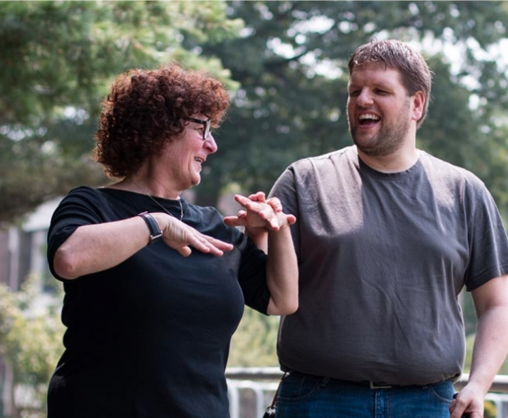 A woman and a man standing outside in front of trees, smiling at each other, and doing tactile sign