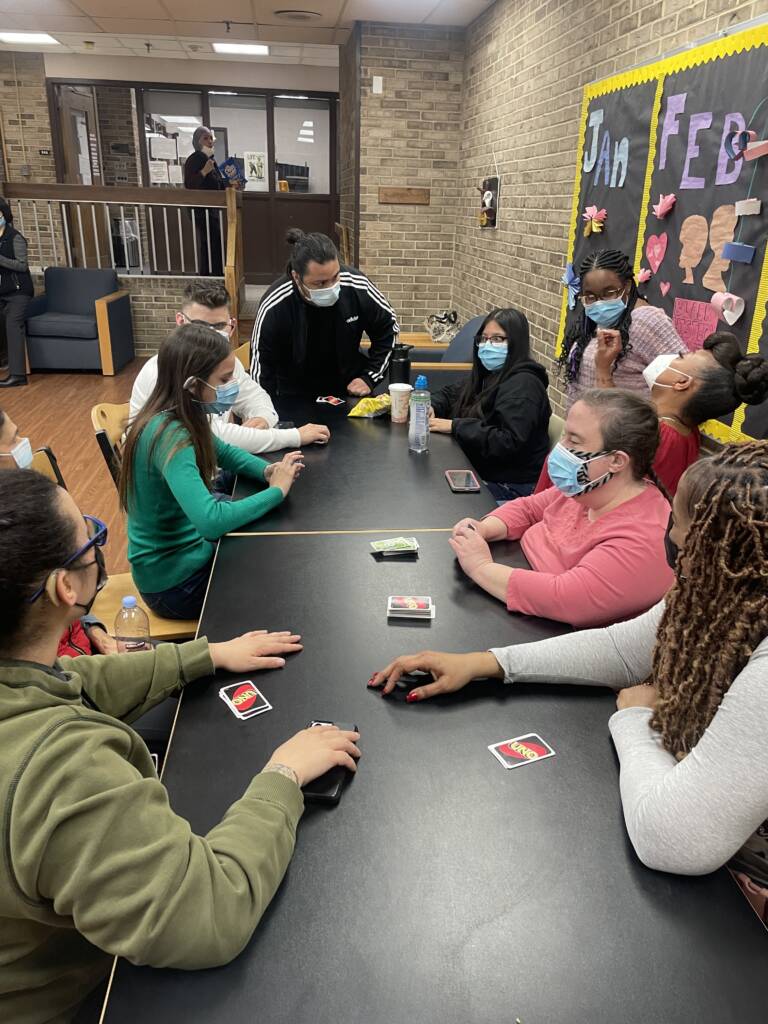 Group of men and women sitting on chairs and standing near table playing Uno in dorm building