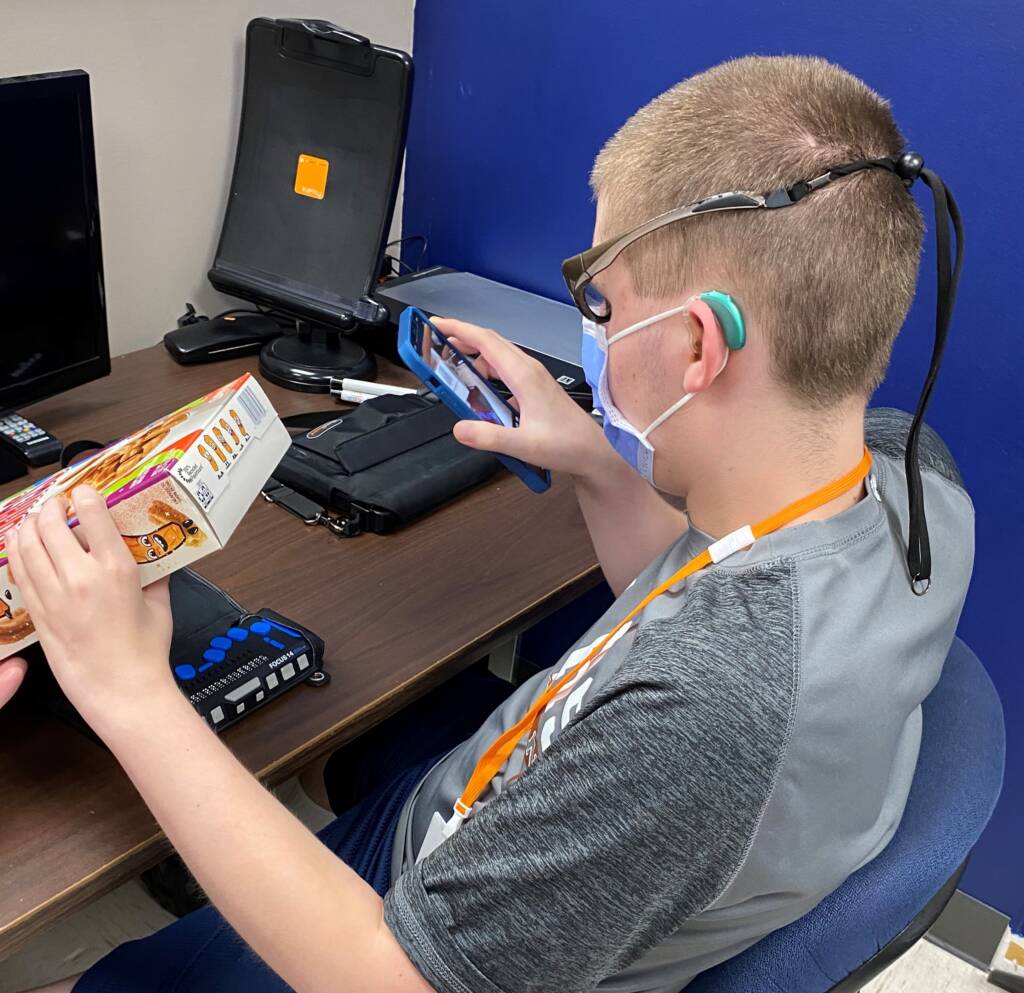 A boy wearing a hearing aid, a face mask, and glasses with a strap taking a photo of the bottom of a cereal box with his phone