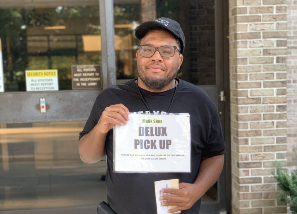 A man holding a sign that says "Delux Pickup - Please tap my shoulder and guide me to the vehicle, I am deaf and low vision"