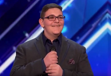 A teenage boy named Christian Guardino smiling, wearing a suit and glasses, and holding a microphone