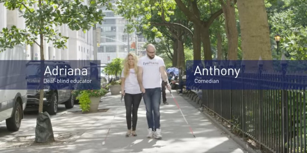 A woman and a man walk on a sidewalk. The man holds a white cane in his left hand.