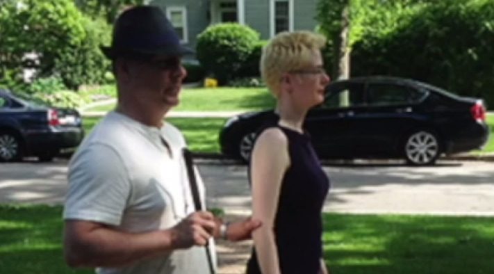 A man holding a white cane while standing next to someone on a sidewalk