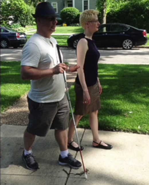 Two people walking on a sidewalk. The man holds a white cane in his right hand and holds the other person’s arm with his left hand