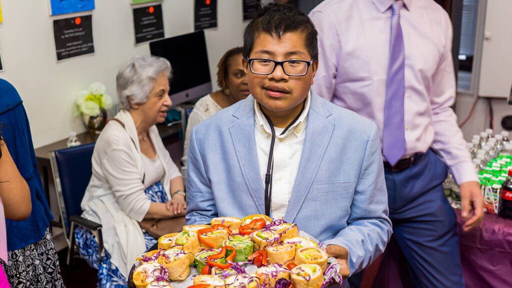 Man looking into the camera, holding a tray of wraps and wearing a suit jacket while surrounded by people in formal attire