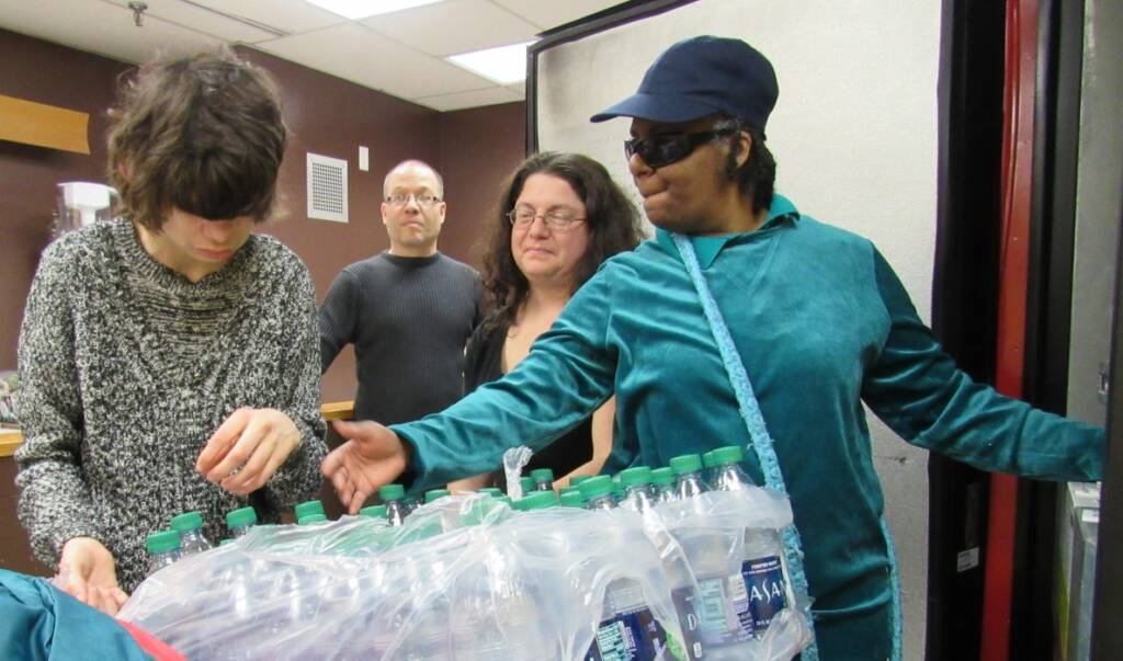 2 people handling a case of Dasani water bottles while 2 staff members wearing black are watching them. One person wears sunglasses