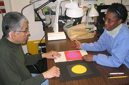 Man with glasses and hearing aid and woman sit across from each other looking at 2 papers with geometric shapes on them