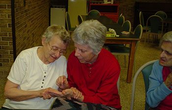 3 older women sitting next to each other. One woman is writing into the palm of another woman with her finger