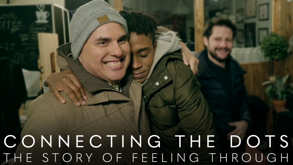 Two men in jackets hugging. Text on bottom of image says “Connecting the Dots: The Story of Feeling Through”