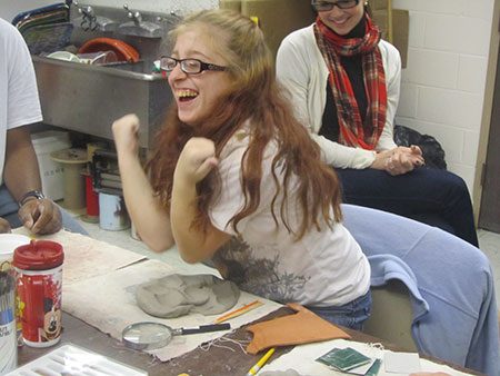 A smiling girl kneading her elbows into clay in an art classroom