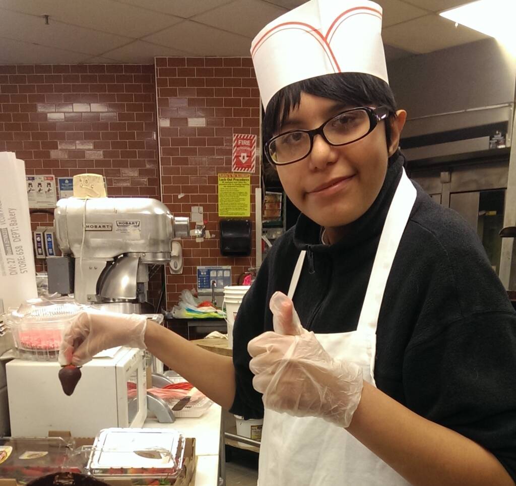 A young person in a kitchen holding a chocolate covered strawberry and wearing a chef hat, apron, and gloves