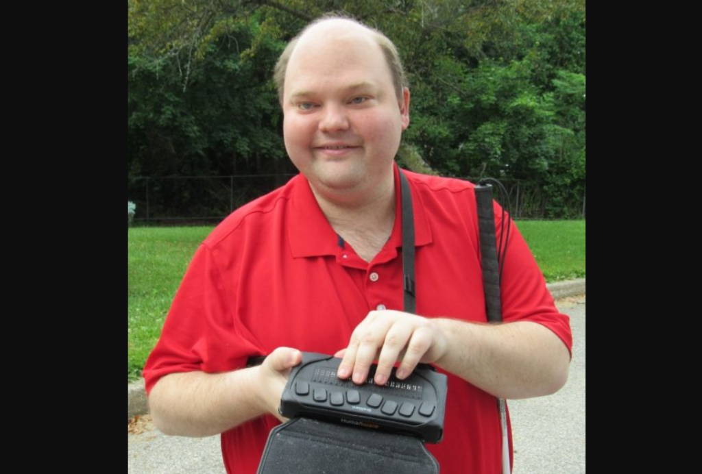 Scott Davert smiling and holding a braille display and white cane outside