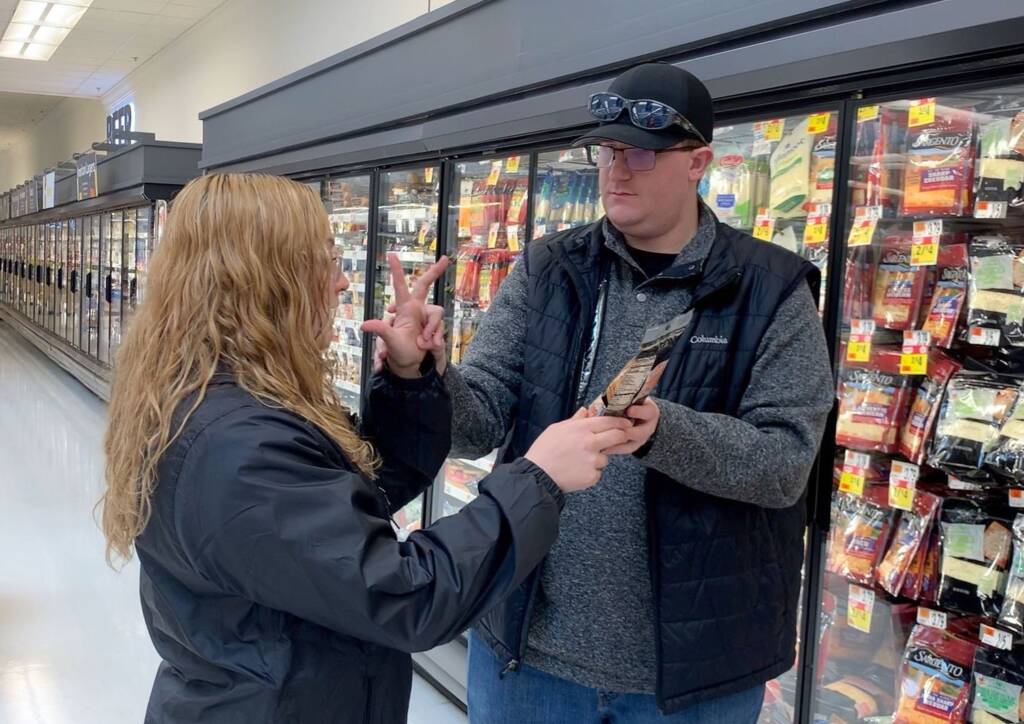 A man holding a bag of cheese and touching the hands of a woman signing. They stand in a refrigerated aisle of a grocery store.