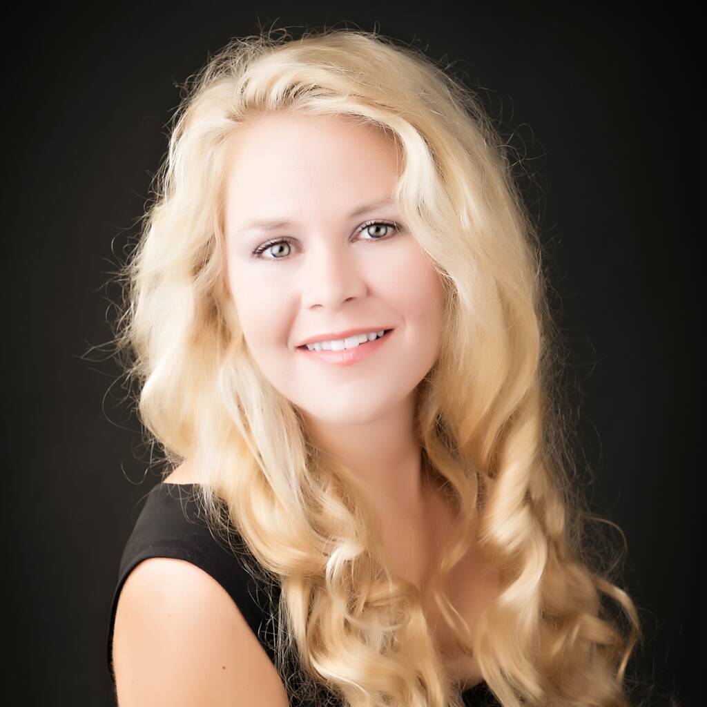 The background is black with a photograph showing, from the top of the head to the waist, of a white woman. She has long curly blonde hair in front of her past her armpits, green/blue eyes, and a smile showing teeth. She is sitting at an angle seen from her right with her head turned and tilted facing the camera. She is wearing a black sleeveless top with a V-neck cut.