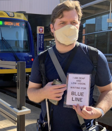 Man standing outside in front of a bus holding a white cane and a sign that says I am deaf and low vision. Waiting for blue line