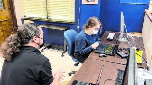 Two people sitting in Helen Keller National Center's technology room. The man looks at the girl who's using a laptop