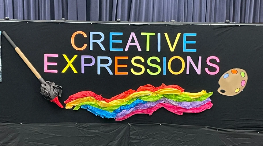 A colorful design made entirely out of paper on a black wall. The words on the design say Creative Expressions, and on the wall is also a large paper paintbrush, a paper swipe of rainbow paint, and a paper paint palette.