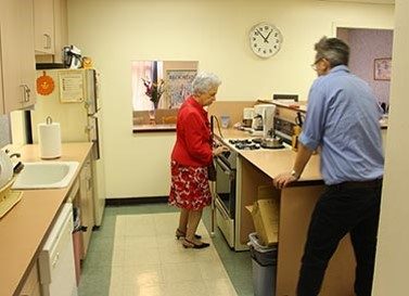 An older woman standing in front of a kitchen stove and holding a white cane. A man stands on the side of the kitchen counter and looks at her
