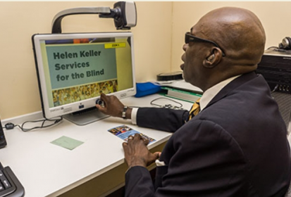 A man using a computer and touching the dial on the screen. The screen says "Helen Keller Services for the Blind" in large text