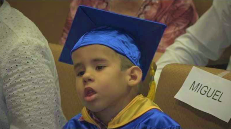 A preschool boy sitting in an auditorium seat wearing a blue graduation cap and gown