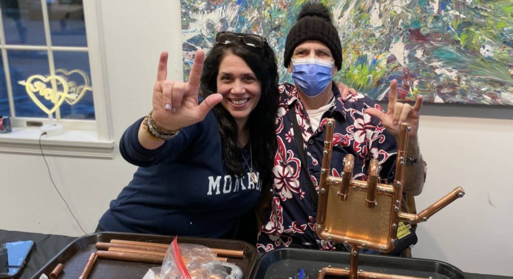 A woman named Jennifer and a male DeafBlind artist named Tony sign I Love You in an art gallery. In front of them is Tony's copper sculpture of a hand signing I Love You