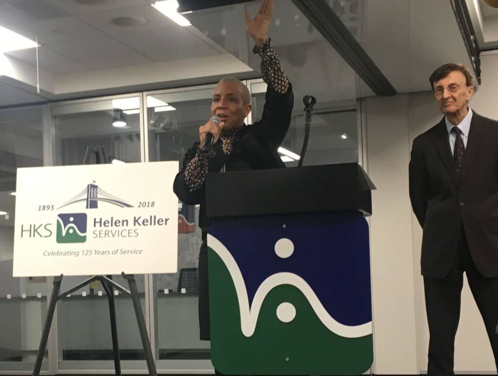 A woman raising her hand next to a man and speaking into a microphone. She is standing behind a podium that has the Helen Keller Services logo on it.