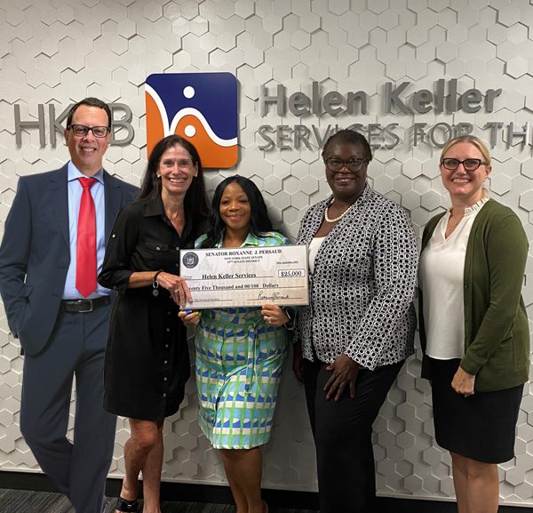 1 man and 4 women standing in front of a Helen Keller Services for the Blind sign. Two of the women are holding a large check made out for $25,000 in front of them.