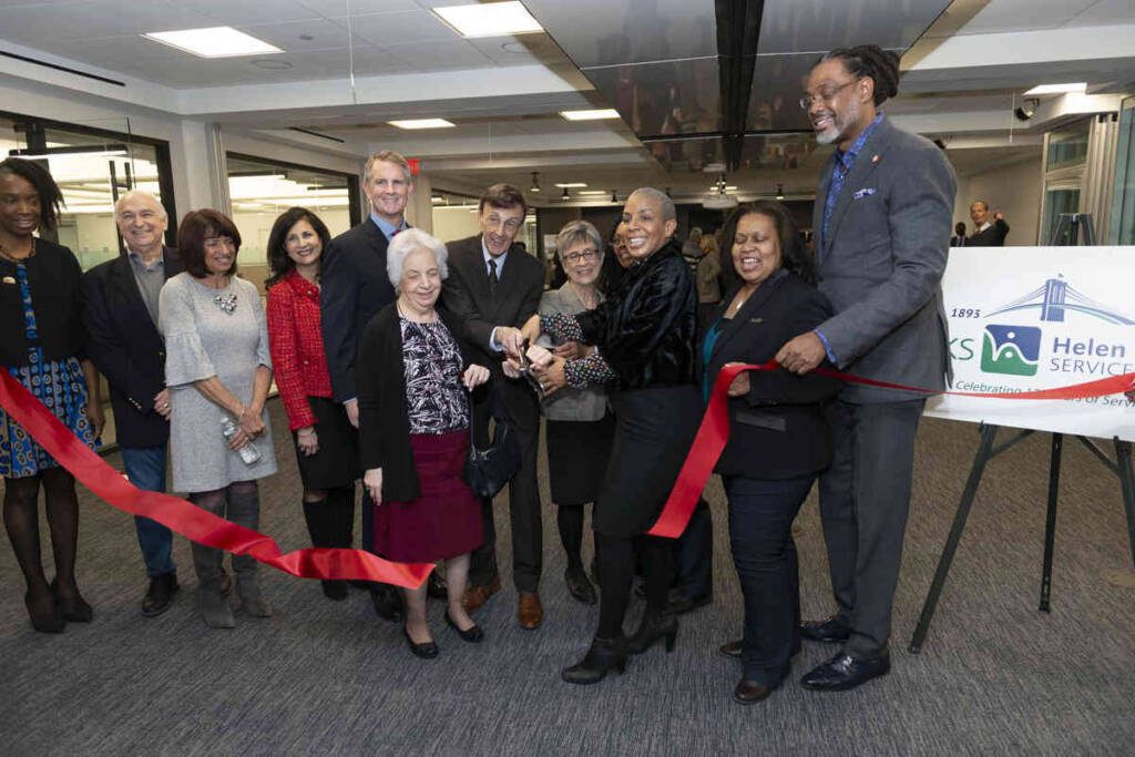 A group of smiling men and women. Many of them hold a scissor together and cut through the middle of a large red horizontal ribbon.