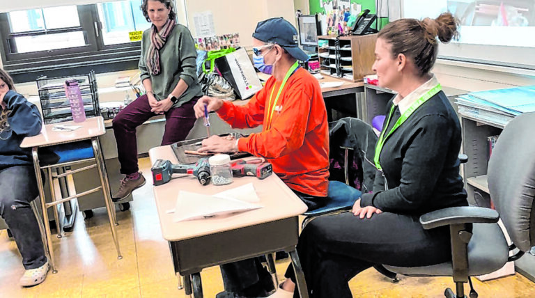 A man holding a screwdriver to copper tubing while sitting at a desk in a classroom. 2 adults and 1 child are shown in the frame watching him assemble his copper sculpture.