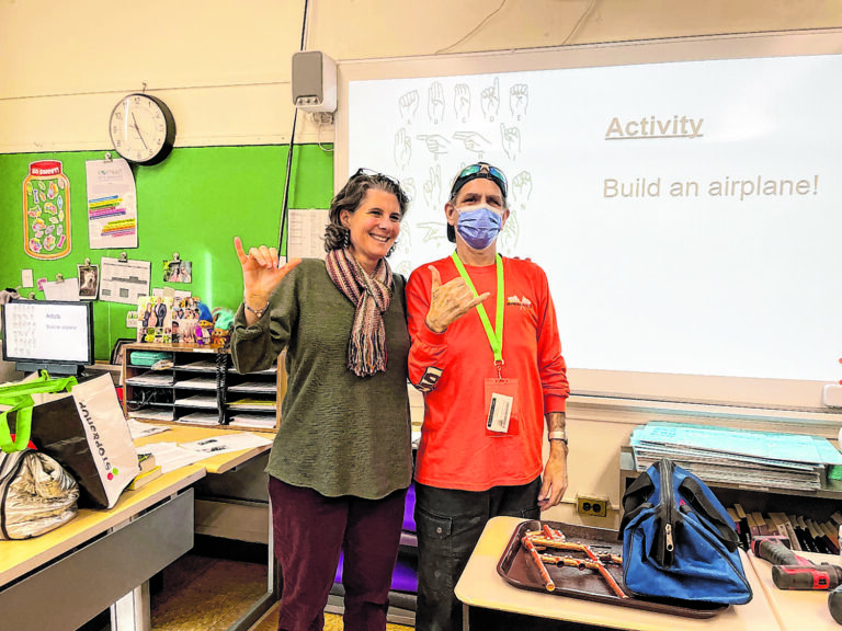 A smiling woman and a man signing I Love You in a classroom. Behind the two people is a large image of the American Sign Language alphabet on a screen.