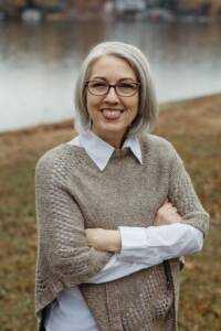 A white woman with straight shoulder-length silver hair standing outside with her arms crossed in front of herself and smiling into the camera outside