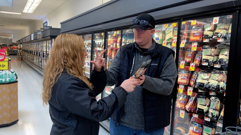 A man and a woman touching hands and holding a bag of cheese in a grocery store aisle that has cheeses in the fridges behind them