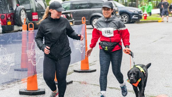 Two women running in the rain. The DeafBlind woman on the right wears a race bib and holds the leash of a black dog guide.
