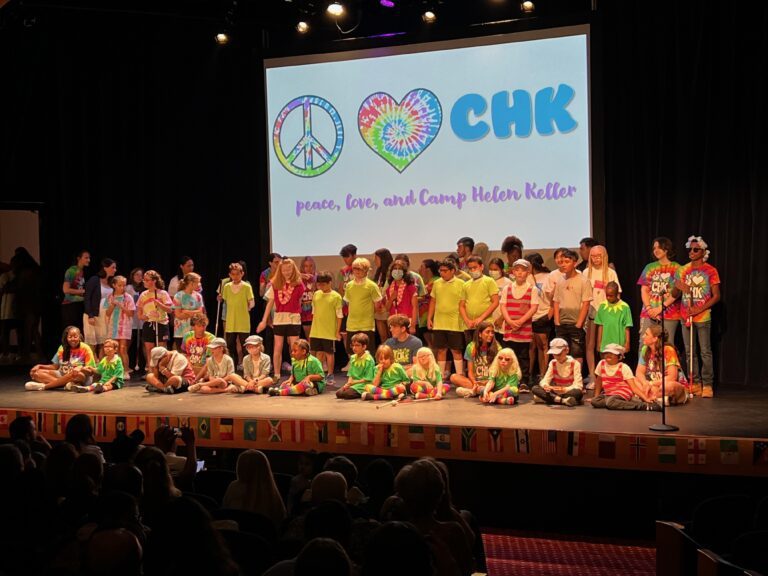 A group of children sitting and standing on a stage wearing green, red, and rainbow tie-dye shirts in front of an audience. Behind the children is a large screen that shows a rainbow peace sign, a rainbow heart, and the acronym "CHK" on it, and underneath those symbols are the words "peace, love, and Camp Helen Keller"