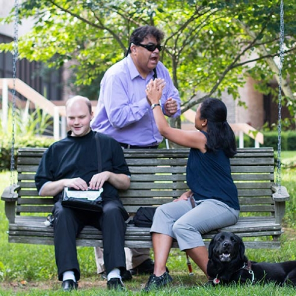 A man sits outside on a bench using a braille display. A woman sits on the bench next to him while touching hands with a man standing behind the bench.