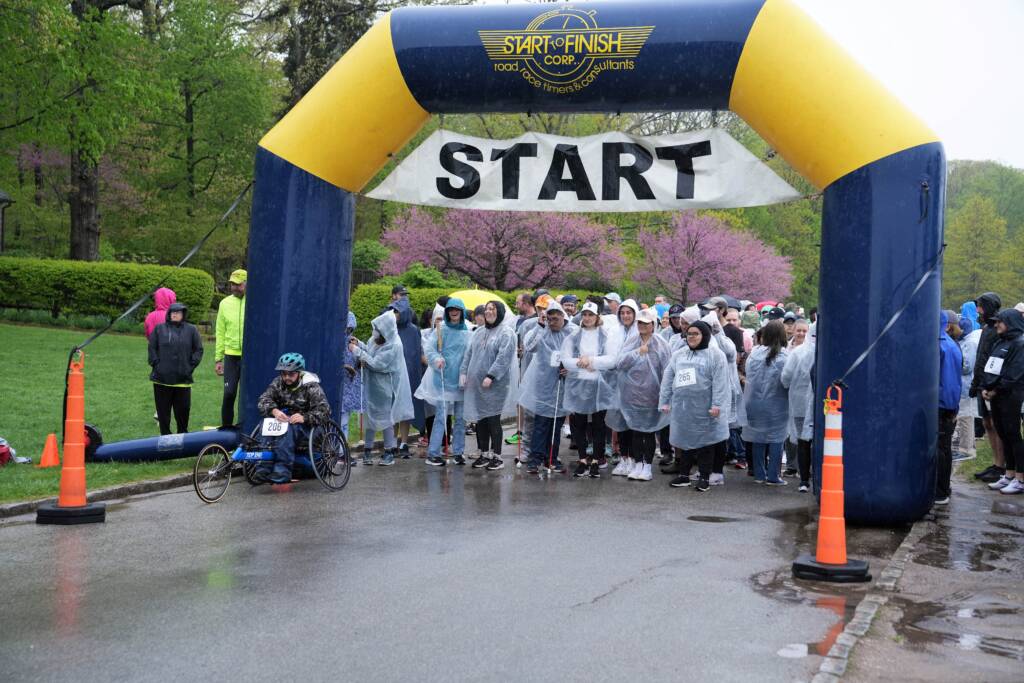 People wearing clear rain ponchos pose outside behind an inflatable race arch that says "Start." Some hold white canes and one person sits in an adapted tricycle.