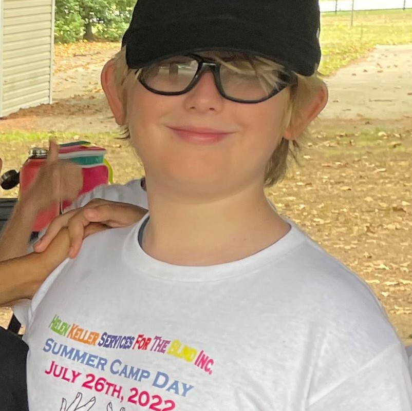 A young boy wearing glasses, a hat, and a Helen Keller Services for the Blind camp t shirt. He smiles into the camera