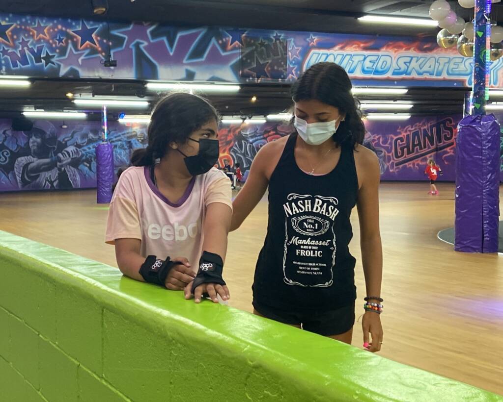 Two girls in a room skating indoors near a green wall. Behind them is a wall full of decorative graffiti.