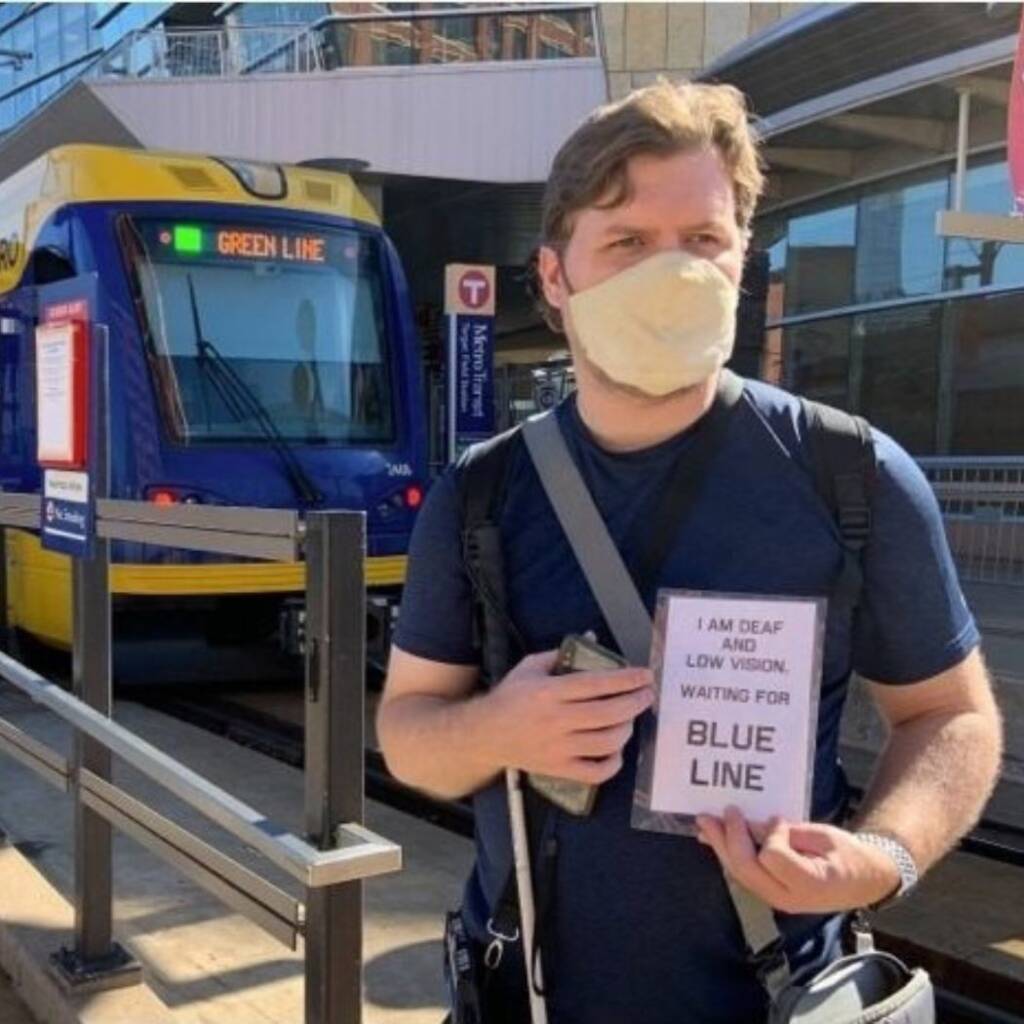 A man who is wearing a COVID-19 mask is standing at a train station, holding a white cane and sign reading: "I am deaf and low vision. Waiting for blue line." He is also wearing a crossbody and holding his cellphone.