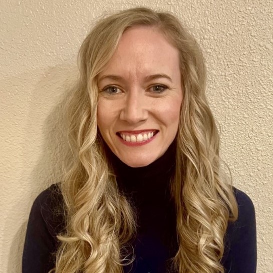 Headshot of a white woman with long, curly blonde hair and blue eyes. She is wearing navy blue turtleneck.