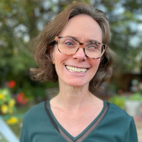 Head shot of a white woman with short brown hair wearing large, dark framed glasses and a v-neck green shirt.