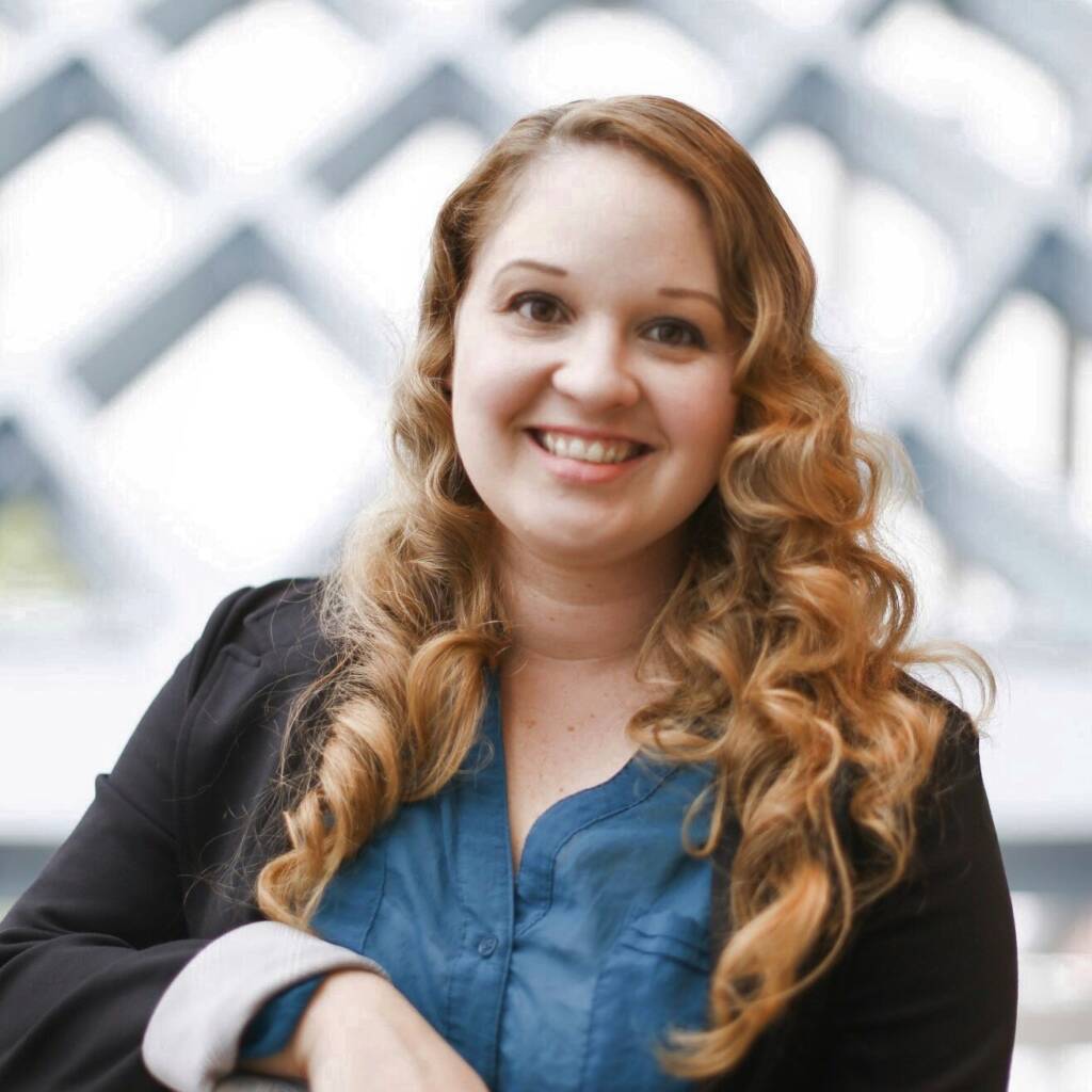 Professional headshot of Tara, a white woman in her early 30s with curly blonde hair and brown eyes smiling in a blazer and button up blouse against an artsy background.