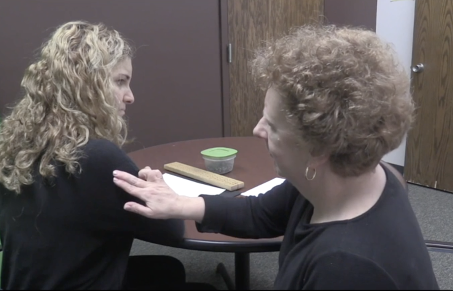 An instructor, a woman with short curly hair, is presenting a haptic signal on the upper arm of a female student with long curly hair who is sitting with her at a round table topped with braille paper, a braille peg board, and a container of pegs.