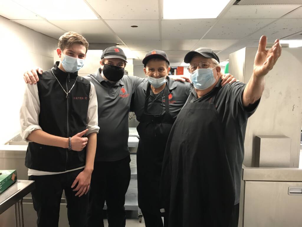 A DeafBlind employee and his work team of 3 other people, wearing face masks and posing for the photo together in the dishwashing area of the cafeteria kitchen.