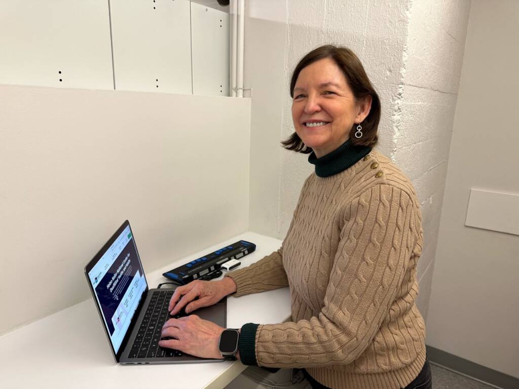 A DeafBlind woman in her 60s smiles while on a laptop that is open to a Helen Keller Services web page. Next to the laptop is a braille display.