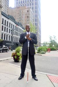 An African-American male wearing a gray sports jacket, with graying hair, and a big smile while proudly holding his white cane in front of tall buildings.