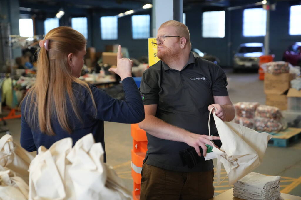 A DeafBlind man works in a warehouse and looks at a woman making an L shape with her hand