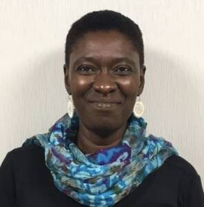 A Black woman with dark chocolate skin, brown eyes, and a short, dark brown natural hairstyle. She is wearing a bright blue scarf over a black shirt, looking at the camera and smiling