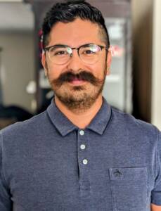 A Latino male smiling and wearing eyeglasses. He is wearing a navy polo shirt with hair combed to the side, has some scruffy hair and a mustache.